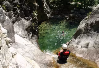 Descente d'un canyon : un pratiquant casqué au bord d'une vasque d'eau claire où nage un autre, entre les rochers