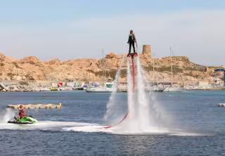 Pratiquant de flyboard propulsé au-dessus de l'eau par deux jets, devant un port et une tour génoise