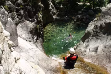 Descente d'un canyon : un pratiquant casqué au bord d'une vasque d'eau claire où nage un autre, entre les rochers
