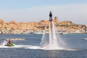 Pratiquant de flyboard propulsé au-dessus de l'eau par deux jets, devant un port et une tour génoise